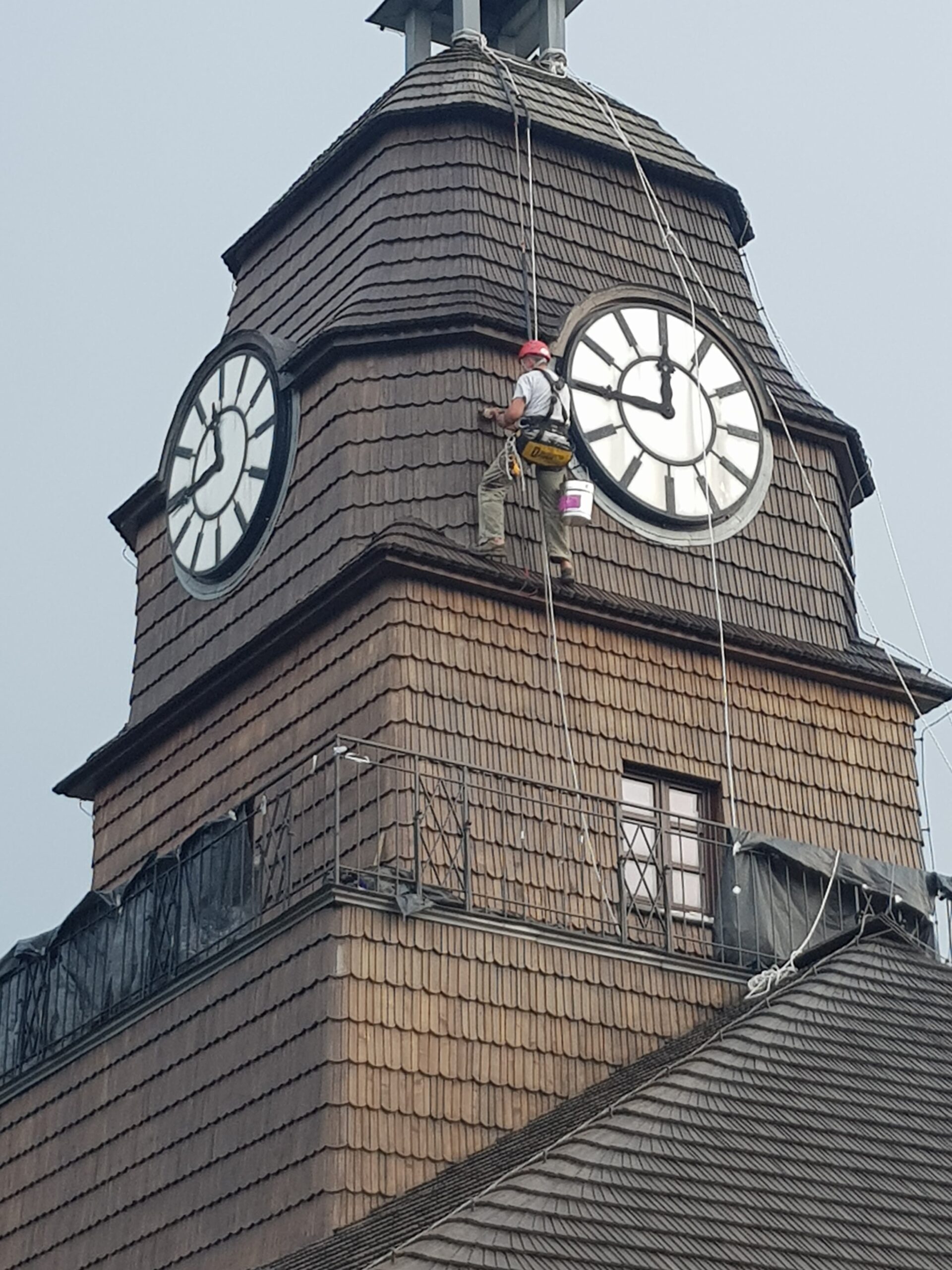 Alpinista przemysłowy, wisząc na linie, maluje wieżę zabytkowej Huty Uthemanna w Katowicach. Z każdej strony wieży znajduje się wielki zegar.