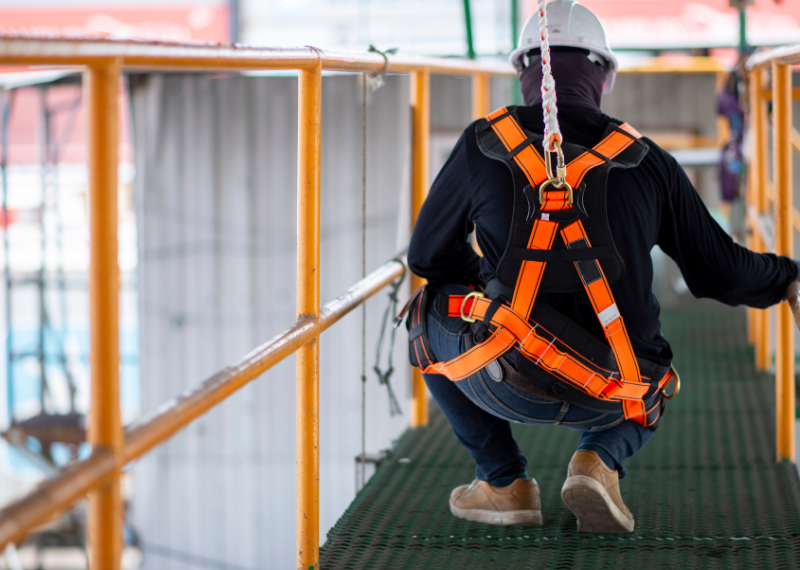 Mężczyzna w pomarańczowych szelkach bezpieczeństwa kuca na mostku w zakładzie przemysłowym.
