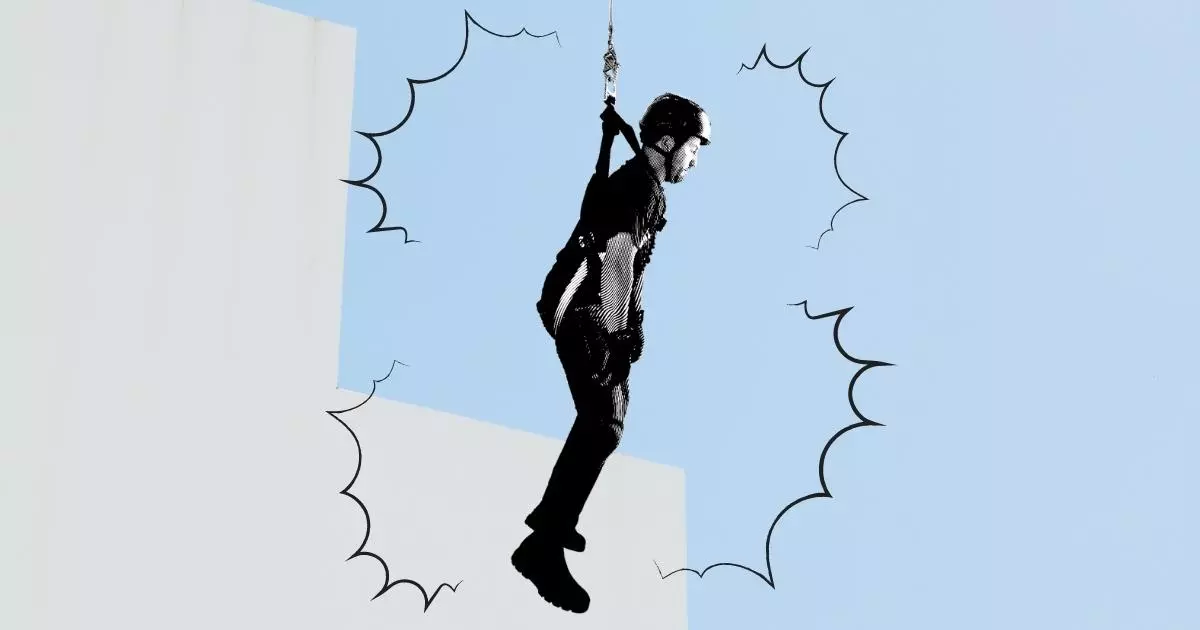 Mężczyzna w szelkach bezpieczeństwa wisi bezwiednie na linie. W tle szary budynek i błękitne niebo.