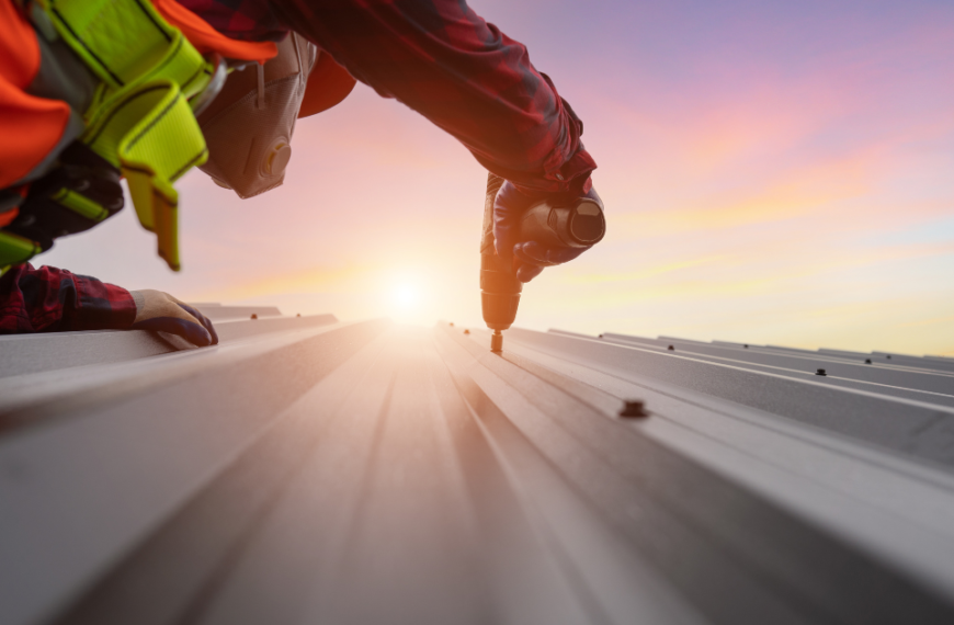 Mężczyzna w roboczym ubraniu przykręca wkręt do blachy na dachu o zachodzie słońca.